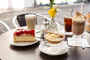Cafétisch im Außenbereich mit ein Stück Erdbeerkuchen, Eiskaffee, Affogato, Eistee, Cafe Latte und einer kleinen Tischvase.