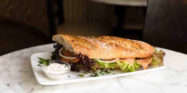 Belegtes Baguette mit Räucherlachs, Salat und Zwiebelringen auf einem länglichen Teller neben einem kleinen Schälchen Dip.