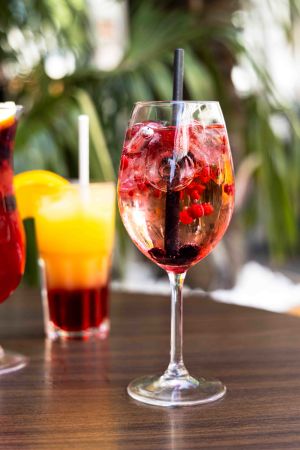 Cocktail im Weinglas mit Eiswürfeln, Beeren und Strohhalm, im Hintergrund weitere bunte Getränke.