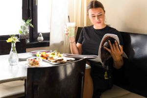 Frau sitzt an einem Cafétisch, liest ein Magazin und hält ein Glas Matcha Latte. Auf dem Tisch stehen Speisen und eine Blume.