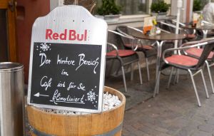 Aufsteller mit Red-Bull-Logo auf einer Pflanzkufe vor Cafétischen. Text weist auf klimatisierten Bereich hin.