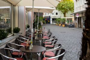 Bestuhlte Terrasse eines Cafés in einer Pflasterstraße mit Sonnenschirmen und kleinen Tischvasen.