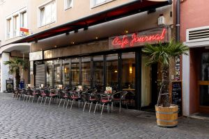 Außenansicht des Café Journal mit roter Leuchtschrift, gefalteter Glasfront, Bestuhlung und Palme im Holzfass.