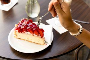Hand führt eine Kuchengabel in ein Stück Erdbeerkuchen auf einem weißen Teller auf einem Holztisch.