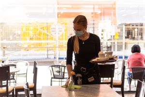 Junge Kellnerin mit Gesichtsmaske räumt leere Kaffeetassen ab und wischt über den abgeräumten Tisch