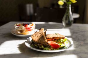 Teller mit einem halbierten Vollkorn-Sandwich, Tomate und Salatgurke neben einer Schale mit Obstsalat auf einem Marmortisch.