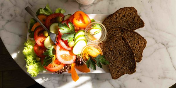 Draufsicht auf einen Teller mit buntem Gemüsesalat, Dips und drei Scheiben Vollkornbrot auf einem Marmortisch.