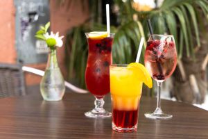 Drei verschiedene farbenfrohe Cocktails und Mocktails mit Strohhalmen auf einem dunklen Holztisch.