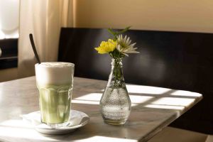 Hohes Glas mit Matcha Latte und Schaumkrone neben einer Glasvase mit gelben und weißen Blumen.