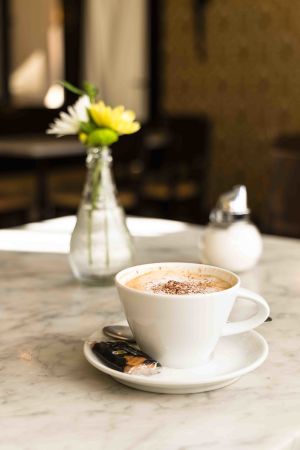 Tasse Cappuccino mit Milchschaum und Kakao auf einem Marmortisch neben einer Blumenvase und einem Zuckerstreuer.